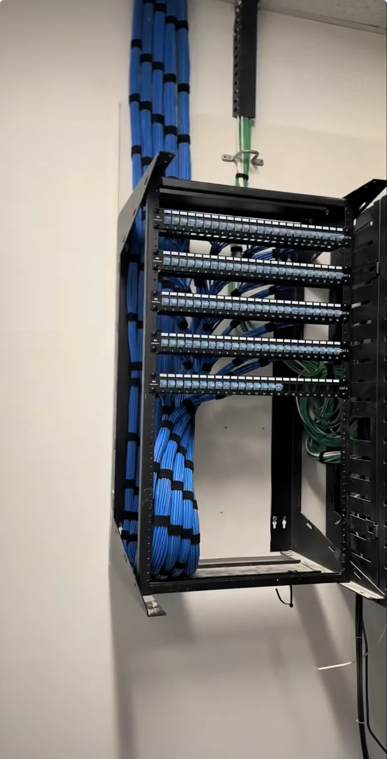 An open wall-mounted equipment rack with five rows of labeled Cat6 patch panels, cabled cleanly with bundled blue cabling running down into the rack.